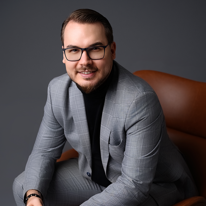 Damian Kowalski ubrany w szary garnitur, siedzi na skórzanym fotelu